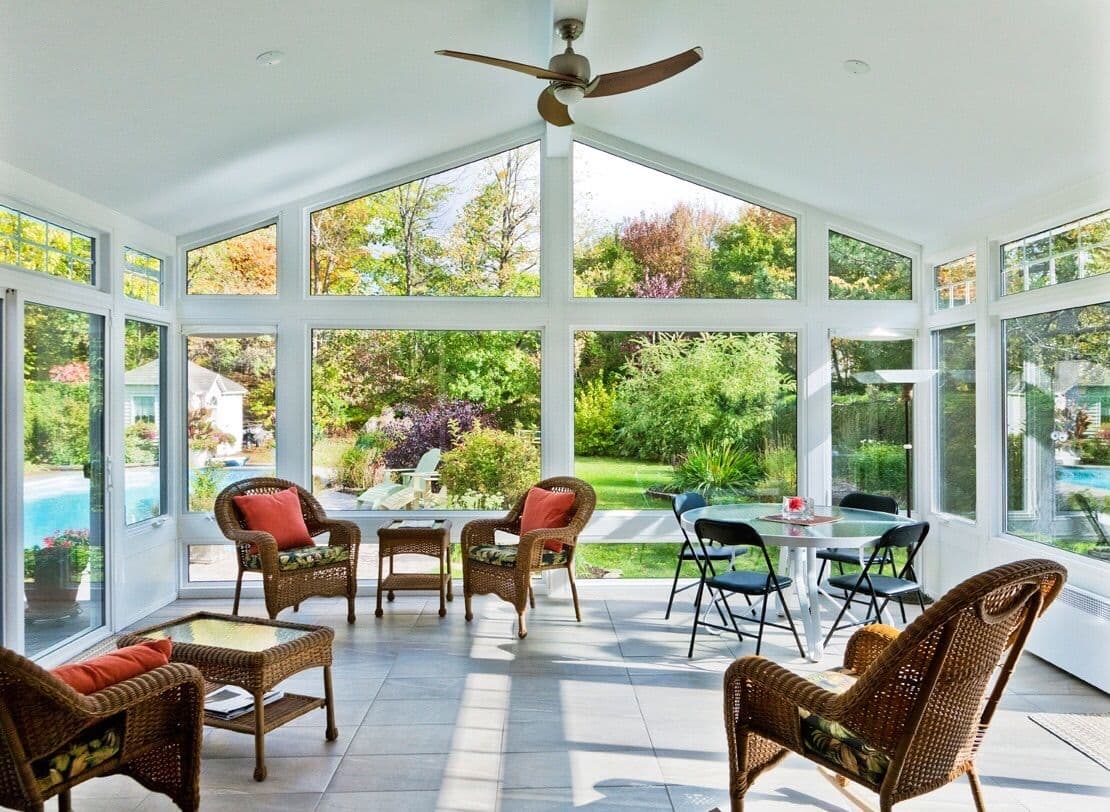 Bright sunroom with large windows overlooking a vibrant garden. Wicker furniture with orange cushions, a ceiling fan, and a round table create a cozy, inviting atmosphere.