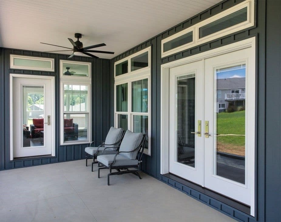 Covered porch with gray siding features white-framed windows and French doors. Two cushioned chairs and a ceiling fan create a relaxed atmosphere.