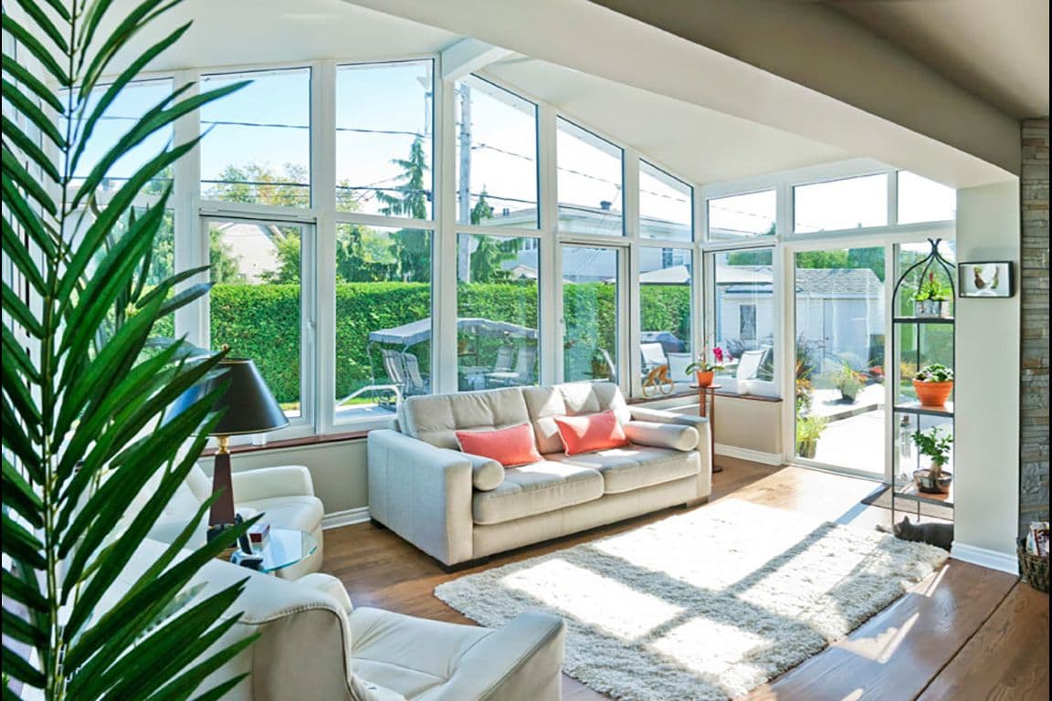 Bright sunlit living room with large windows. Features a cozy sofa with pink cushions, indoor plants, and a fluffy rug on wooden floors, exuding warmth.