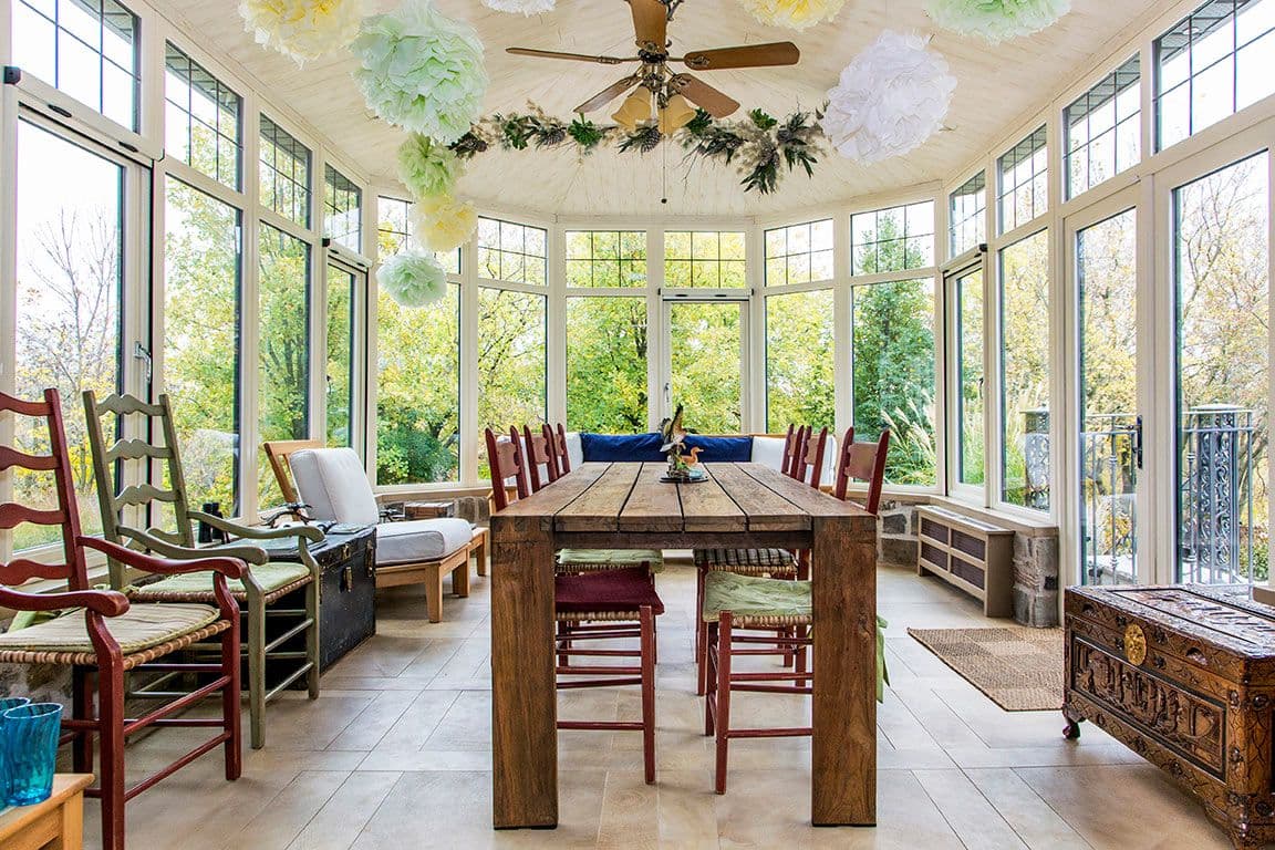 Bright sunroom with large windows, a wooden table, and red chairs. Hanging paper decorations and greenery create a cozy, inviting atmosphere.