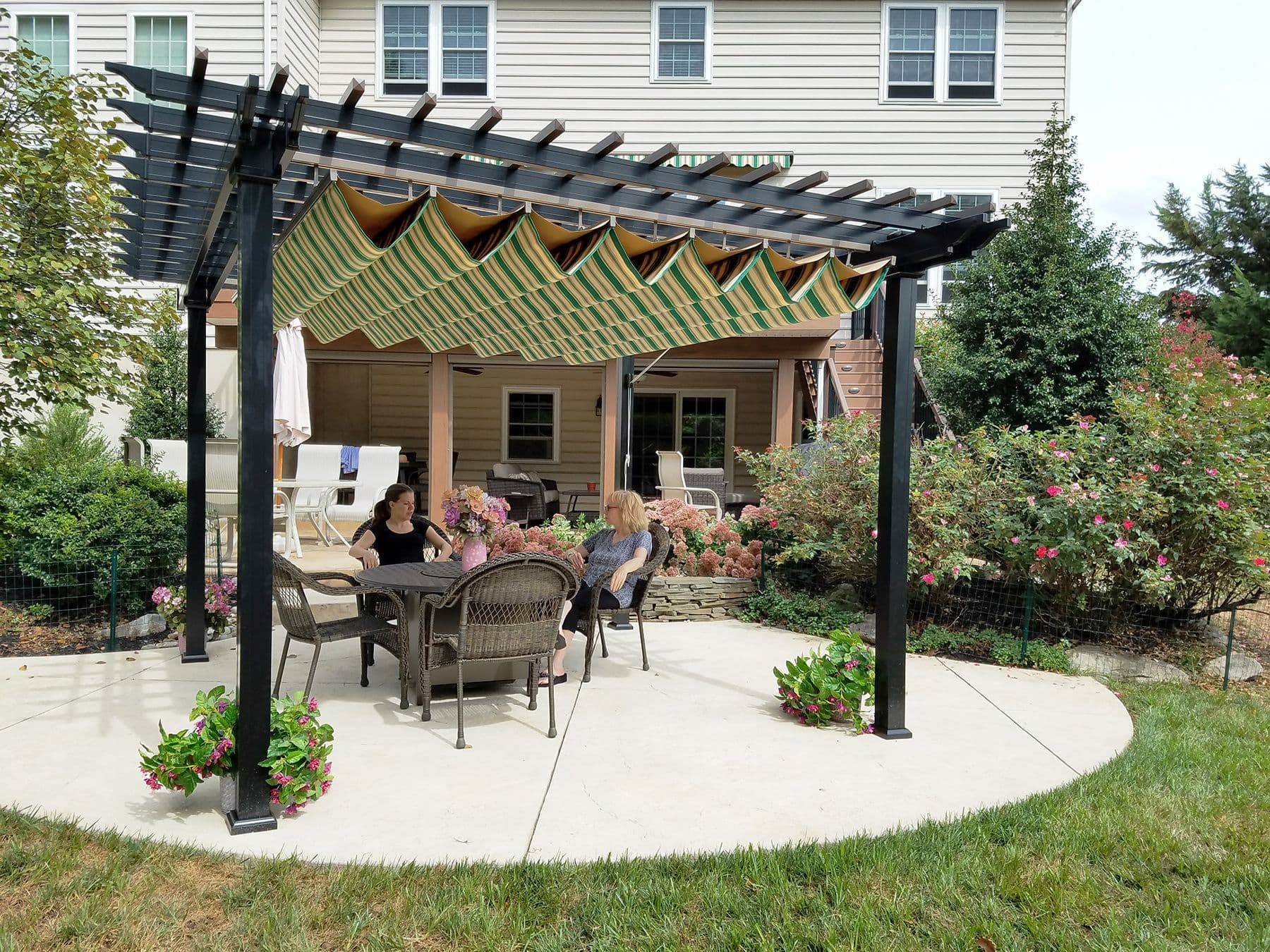 Outdoor patio with two people sitting under a pergola on cushioned chairs. Greenery and flowers surround the area, creating a relaxed atmosphere.