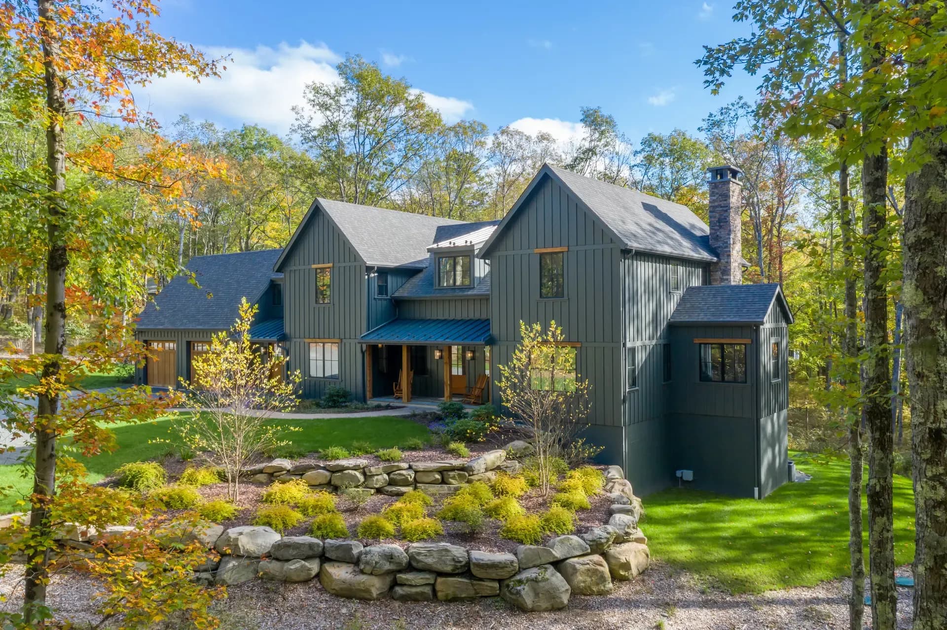Modern, dark grey timber house surrounded by a lush forest with autumn leaves. Stone wall and landscaped garden in the foreground under a clear blue sky.