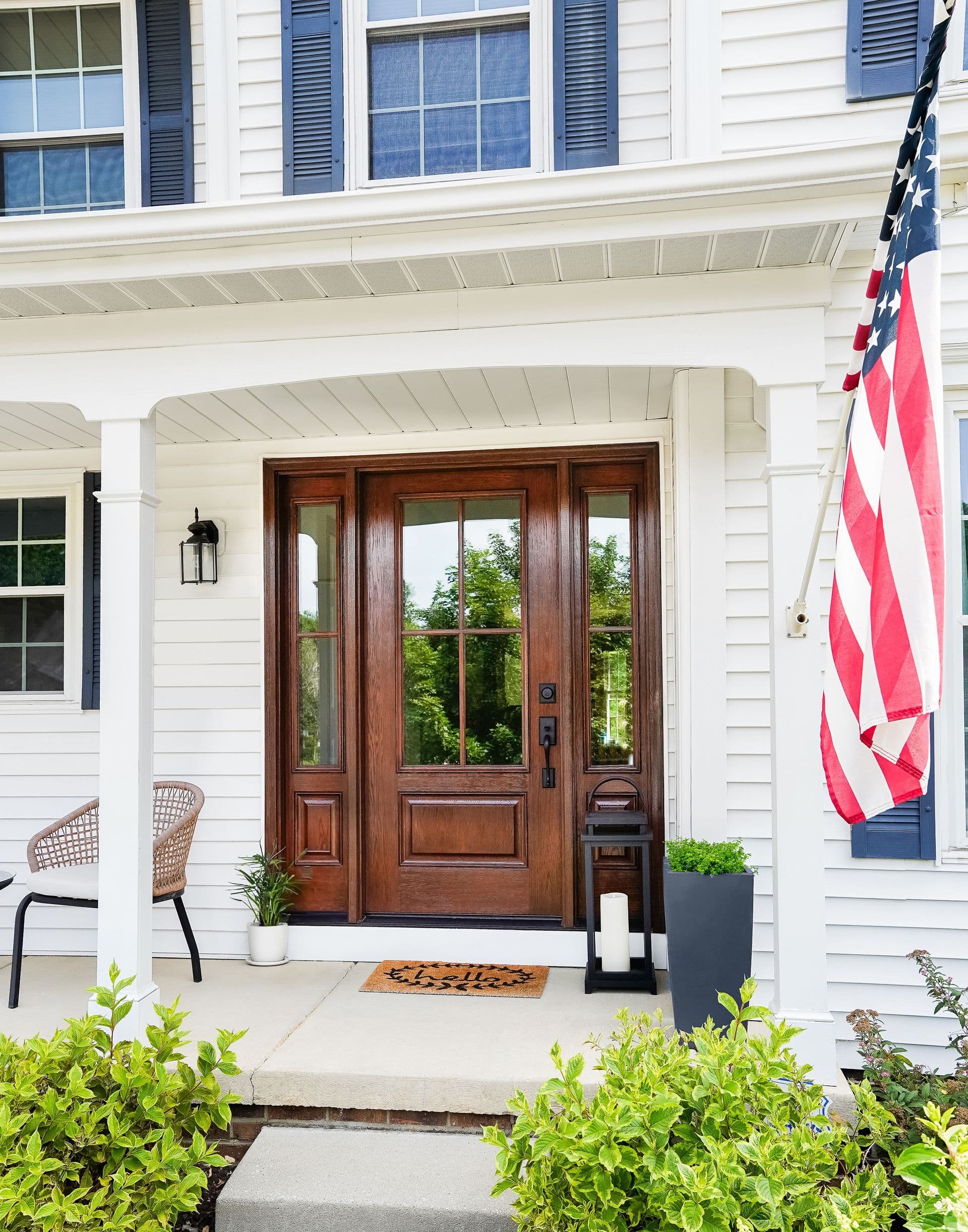 Classic white house entrance with an American flag, wooden door, sidelight windows, potted plants, and a welcoming doormat; evokes a cozy, inviting vibe.