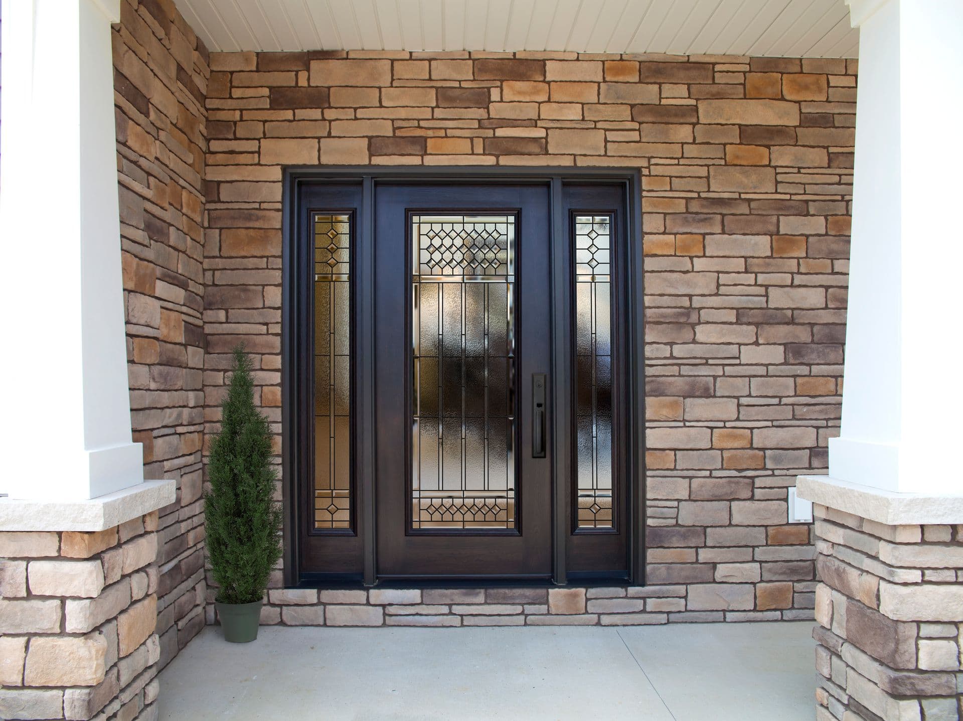 Elegant stone entryway featuring dark wooden door with glass panels and intricate designs, flanked by tall windows. A small potted shrub adds greenery.