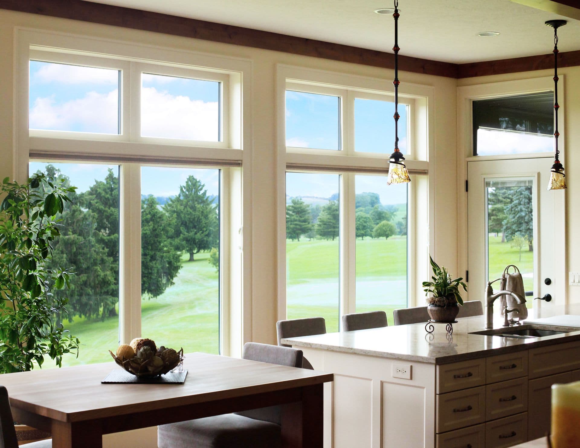 Bright kitchen interior with large windows overlooking a lush green landscape. A wooden table, potted plants, and elegant hanging lights create a serene, inviting atmosphere.