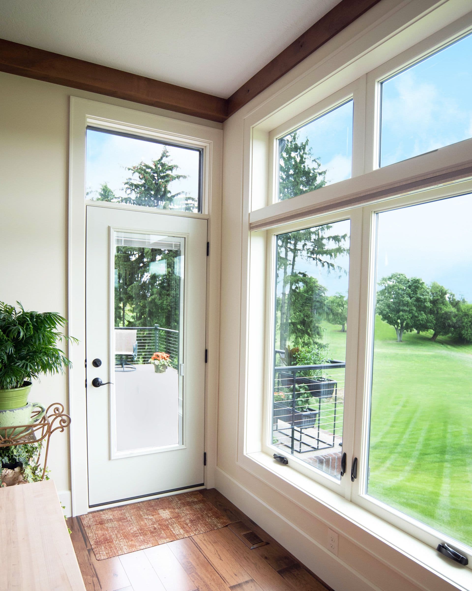 Bright room with large windows showcasing a lush green lawn and trees. A white door with a glass panel leads outside. Indoors, a plant and wooden floor create a serene atmosphere.