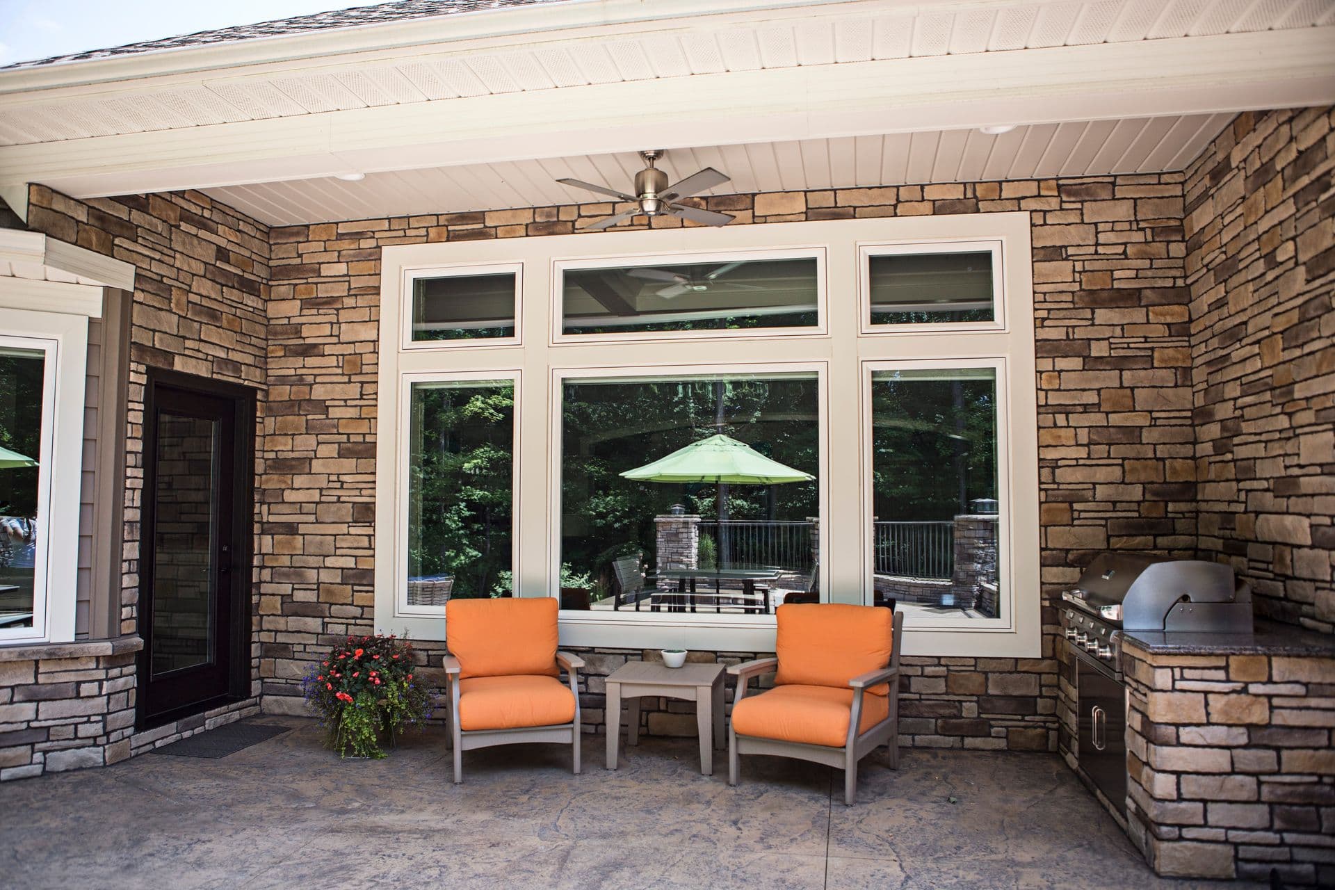 Patio with stone walls, large windows, and two bright orange chairs facing a table. A built-in grill and potted plants add a cozy, inviting feel.
