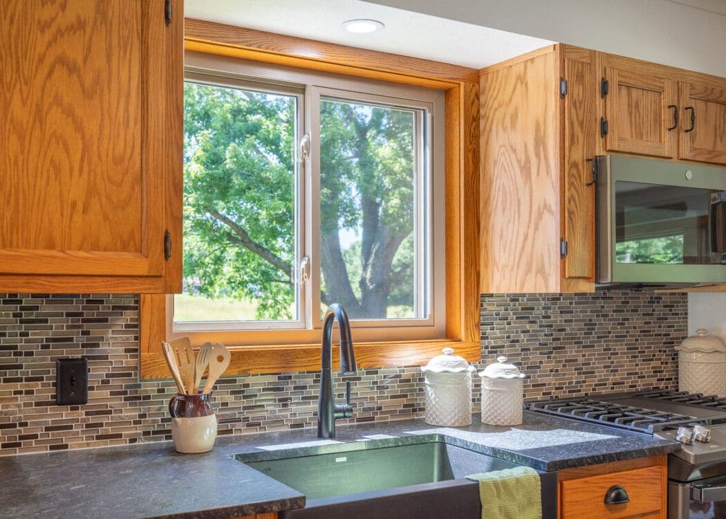 Warm kitchen with wooden cabinets, mosaic tile backsplash, and a large window revealing a leafy tree. Black countertop with a sink, utensils, and jars.