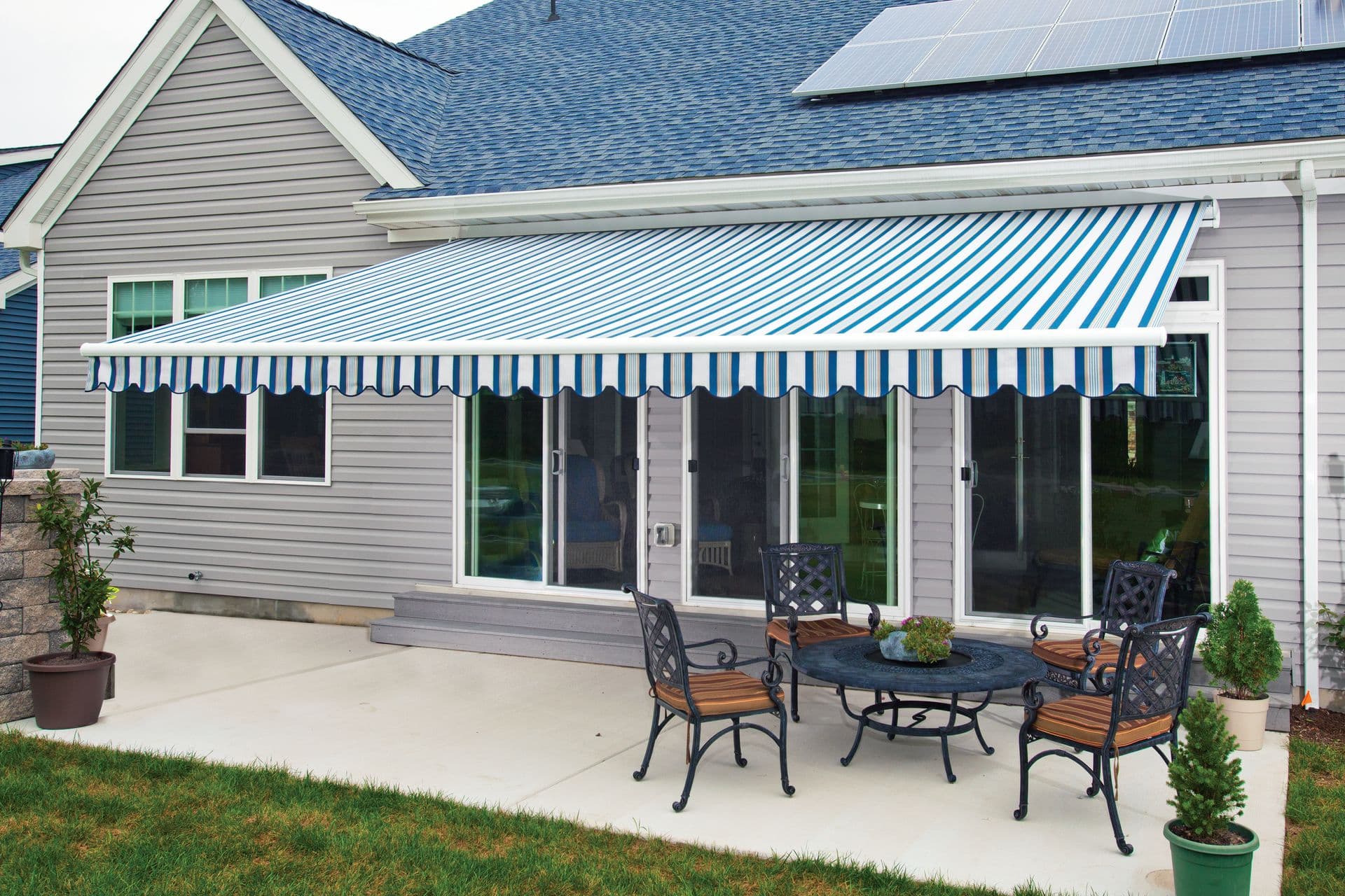 A striped blue and white awning shades a patio with metal furniture. The house exterior is gray with sliding glass doors and solar panels on the roof.