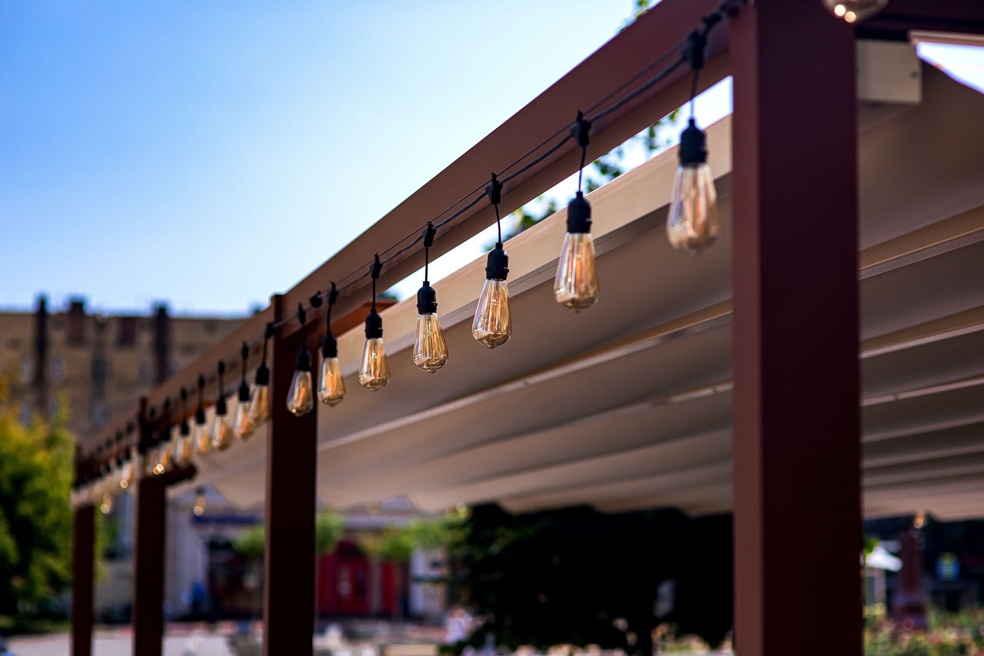 String lights hang from a wooden pergola against a clear blue sky. The background shows blurred buildings and trees, evoking a warm, inviting atmosphere.
