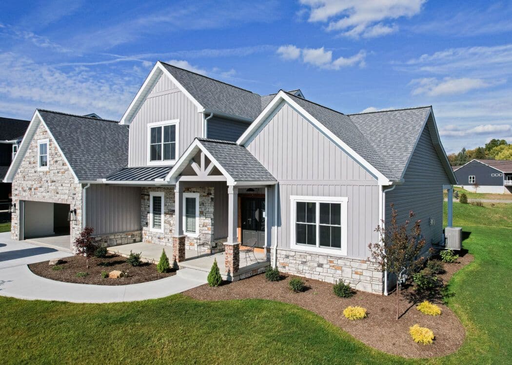 Modern suburban home with gray siding and stone accents, featuring a gabled roof, manicured lawn, and vibrant shrubs under a blue, partly cloudy sky.