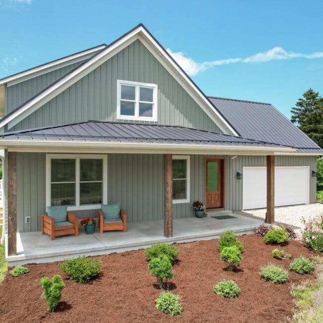 A charming gray house with a steep metal roof, welcoming front porch, and wooden columns. The garden features neatly arranged shrubs, conveying warmth.