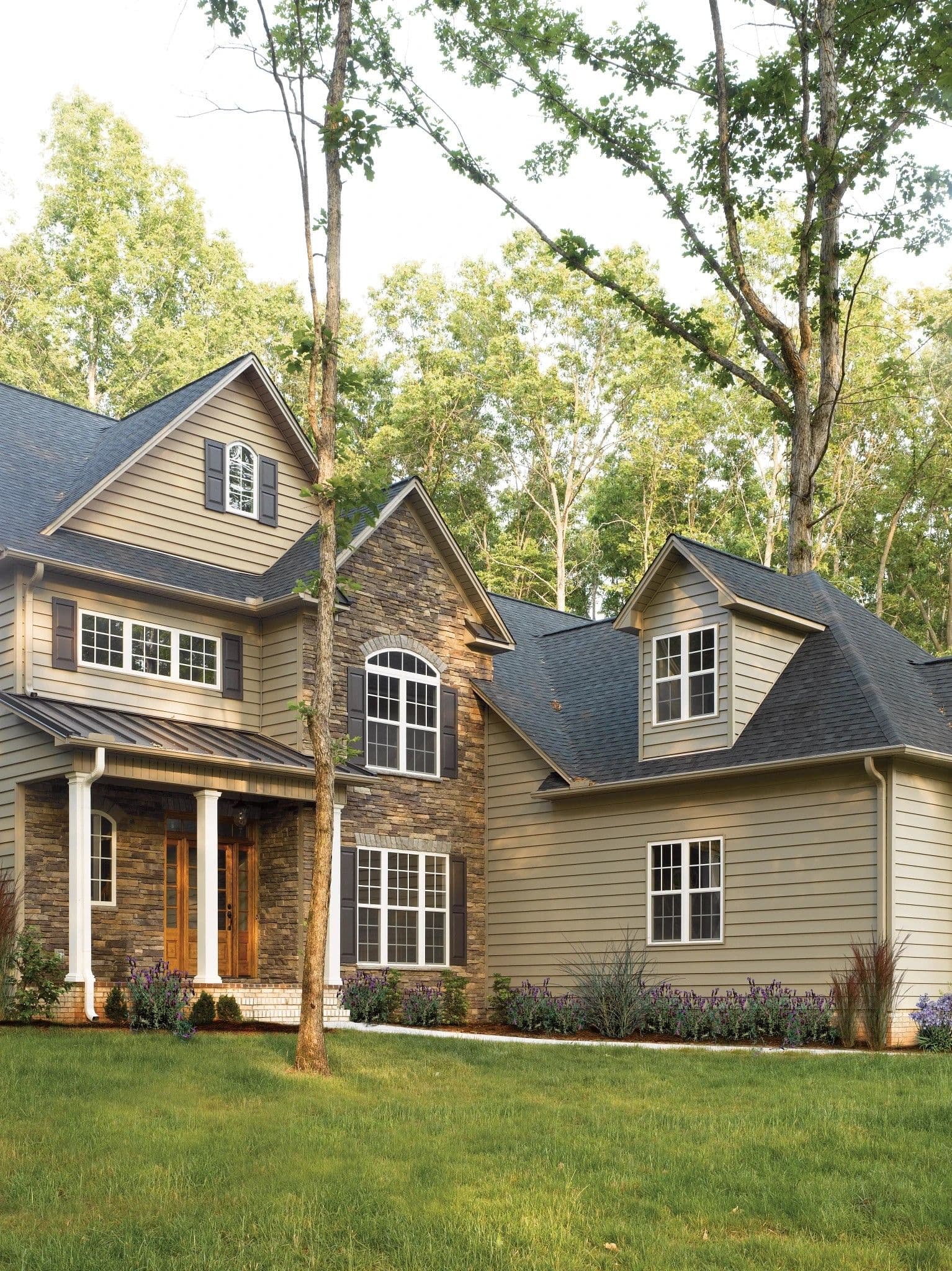 A two-story house with stone and beige siding, surrounded by lush green grass and tall trees, exuding a serene and inviting atmosphere.