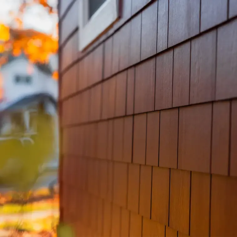 Close-up of a house's brown shingle siding, capturing the texture with sunlight casting a warm glow. Out-of-focus autumn trees are visible in the background.