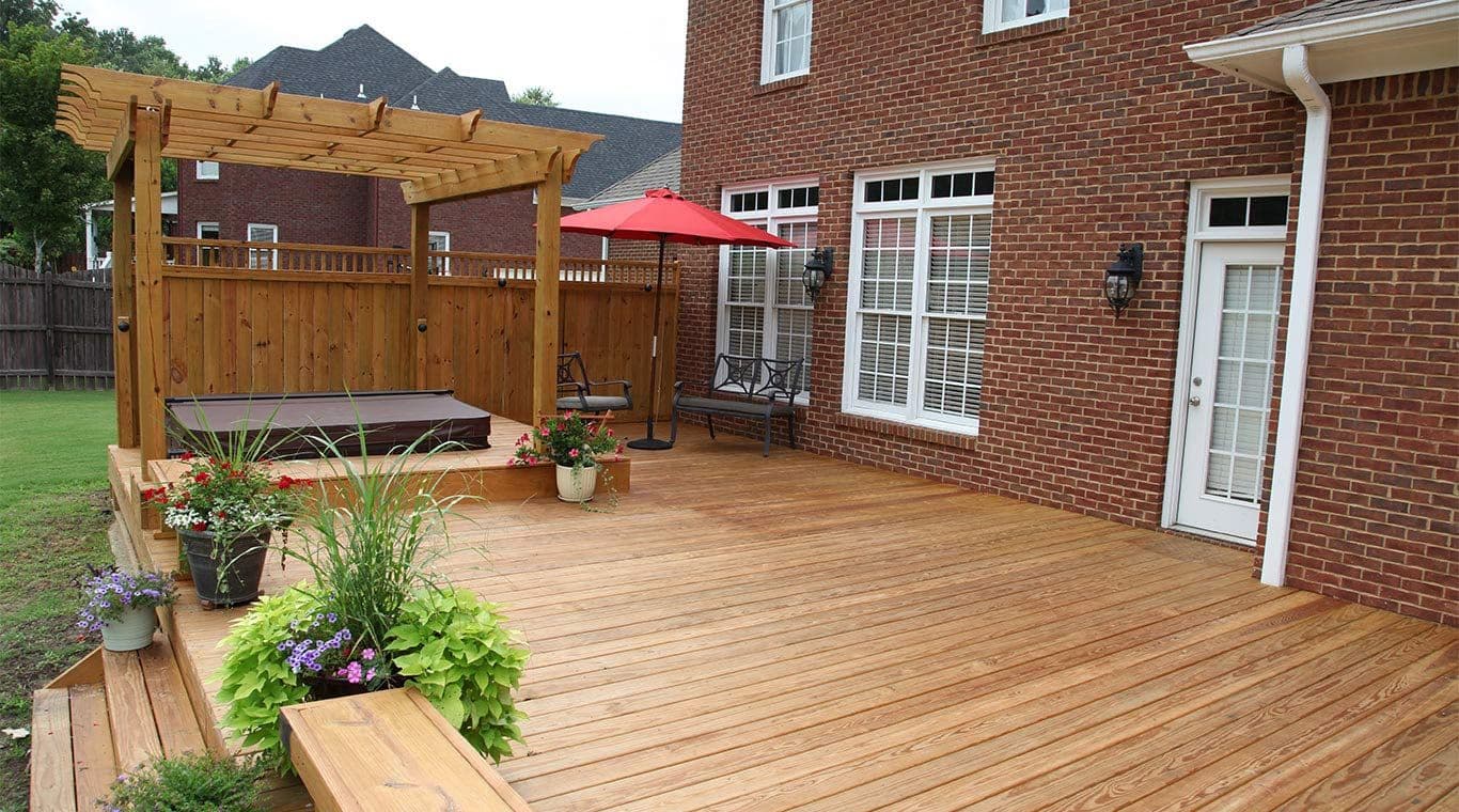 Spacious wooden deck with a pergola and hot tub, potted plants, red umbrella, and brick house walls. Relaxing and inviting outdoor space.