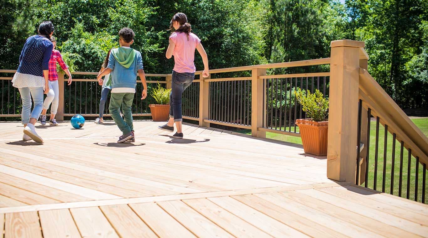 A group of people play soccer on a sunny wooden deck surrounded by lush greenery. Their casual attire and movement convey a cheerful, playful mood.