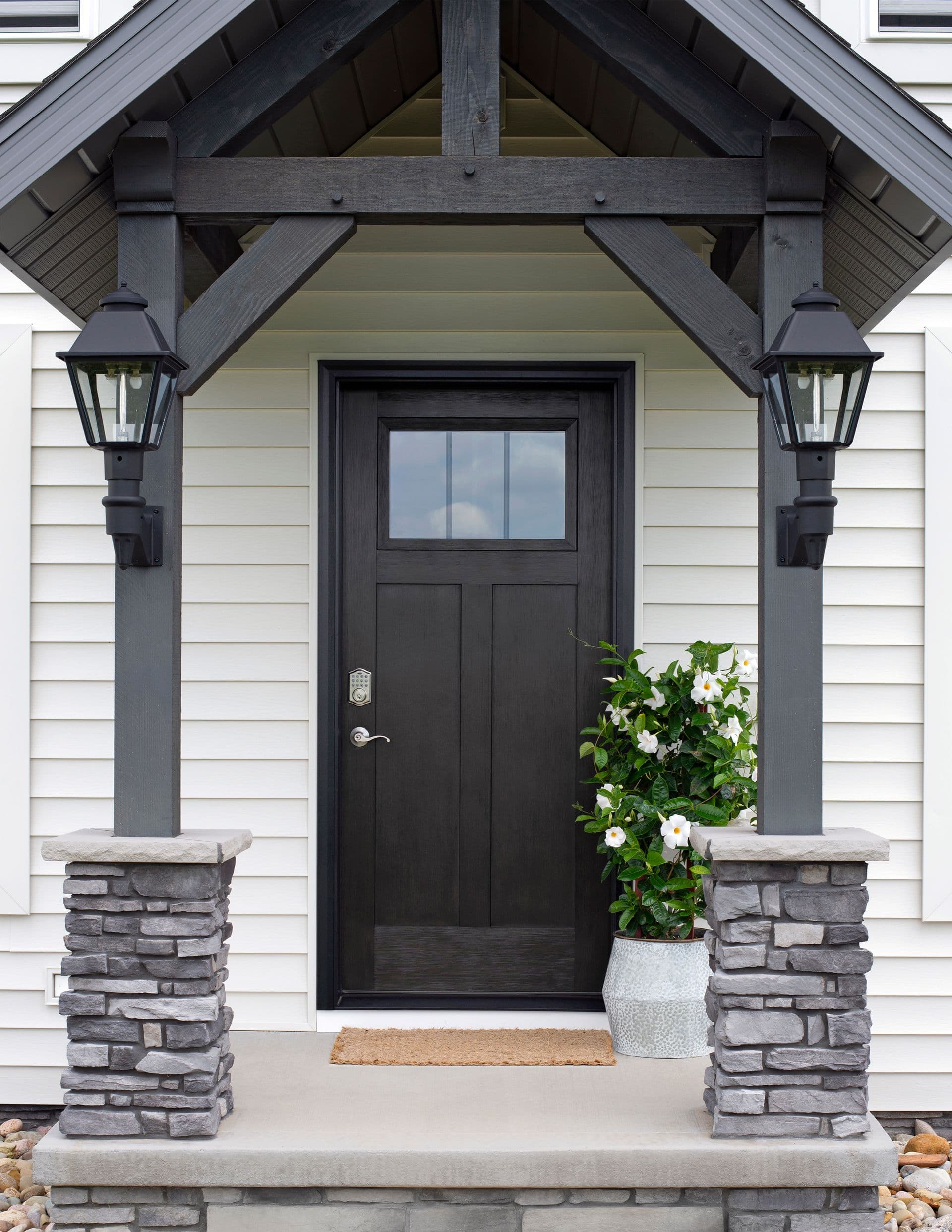 A modern front porch with a black door, stone columns, and white siding. Two lanterns hang beside the door, and a potted plant adds greenery.