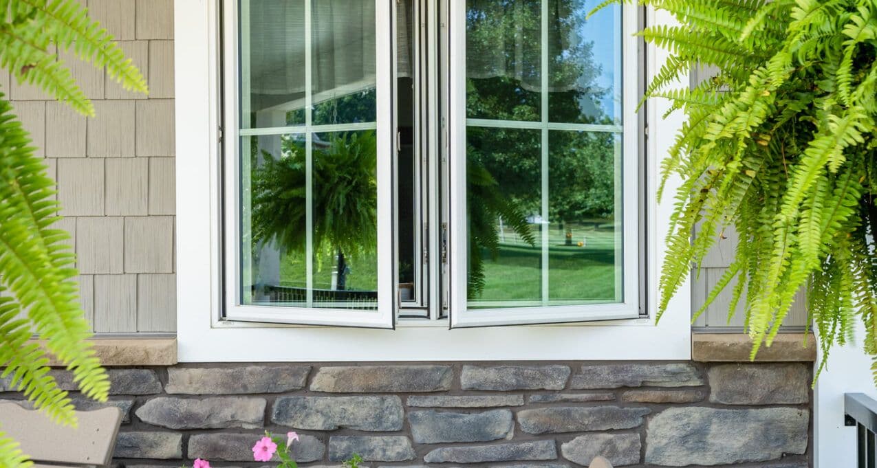 A white-framed window is slightly open on a stone-walled house, reflecting lush green foliage and a sunny lawn, with hanging ferns adding a serene feel.