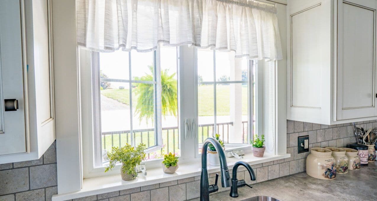 Bright kitchen scene with a window view of a sunny garden. Green plants line the windowsill, and a sleek black faucet is centered below. Calm and inviting.