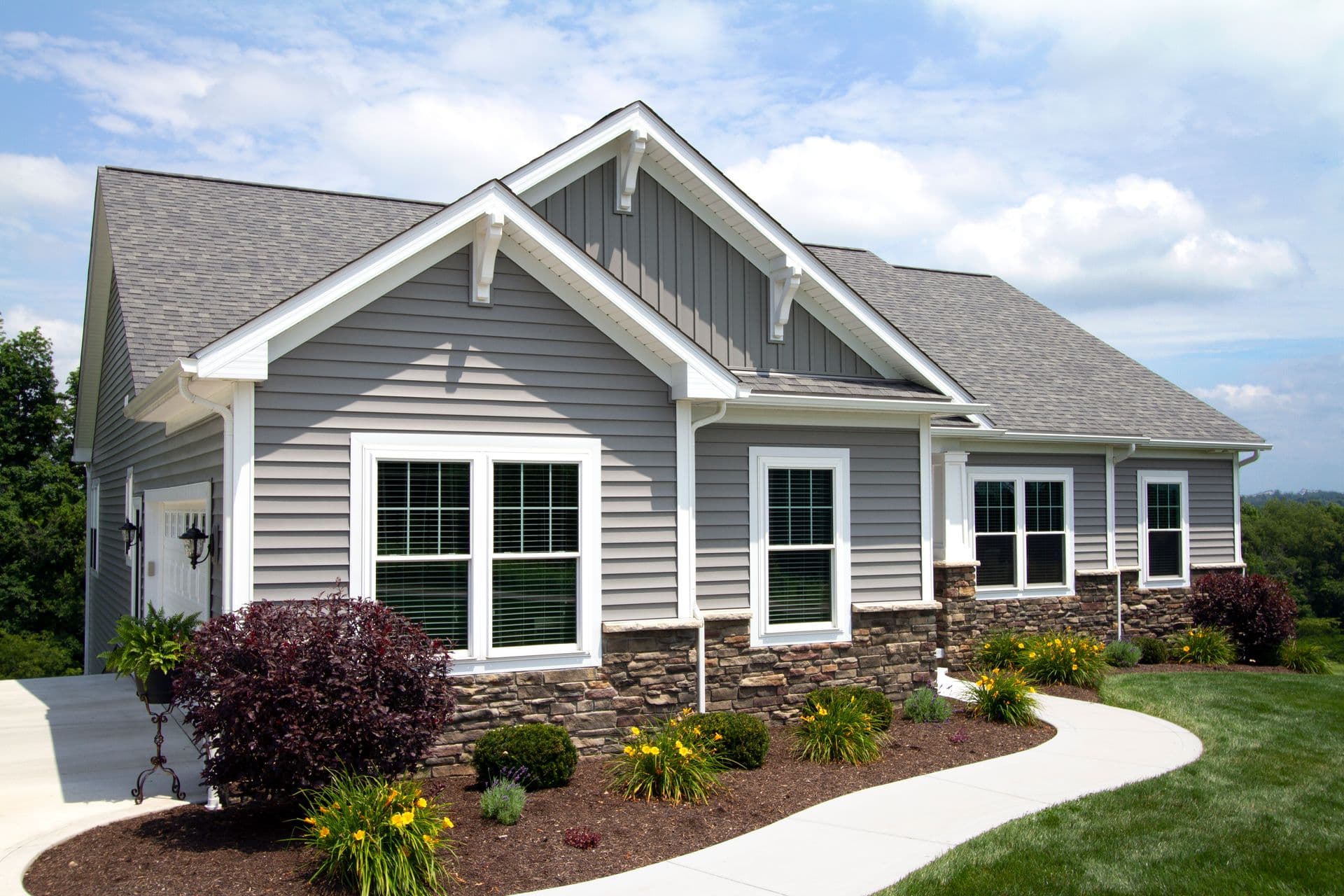 Modern suburban home with gray siding, stone accents, and a sloped roof. Lush landscaping features green shrubs and yellow flowers along a curved walkway.