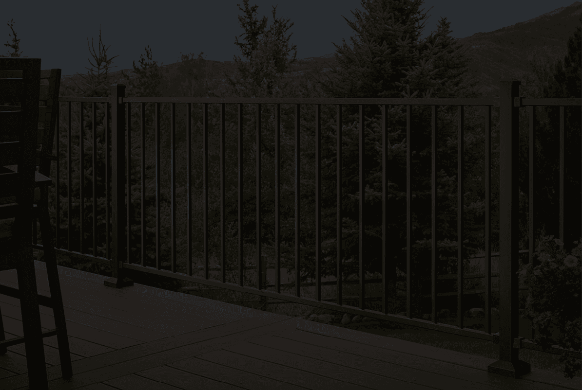 A wooden deck with dark metal railing overlooks a lush landscape with evergreen trees and mountains. A chair is partially visible, suggesting relaxation and serenity.