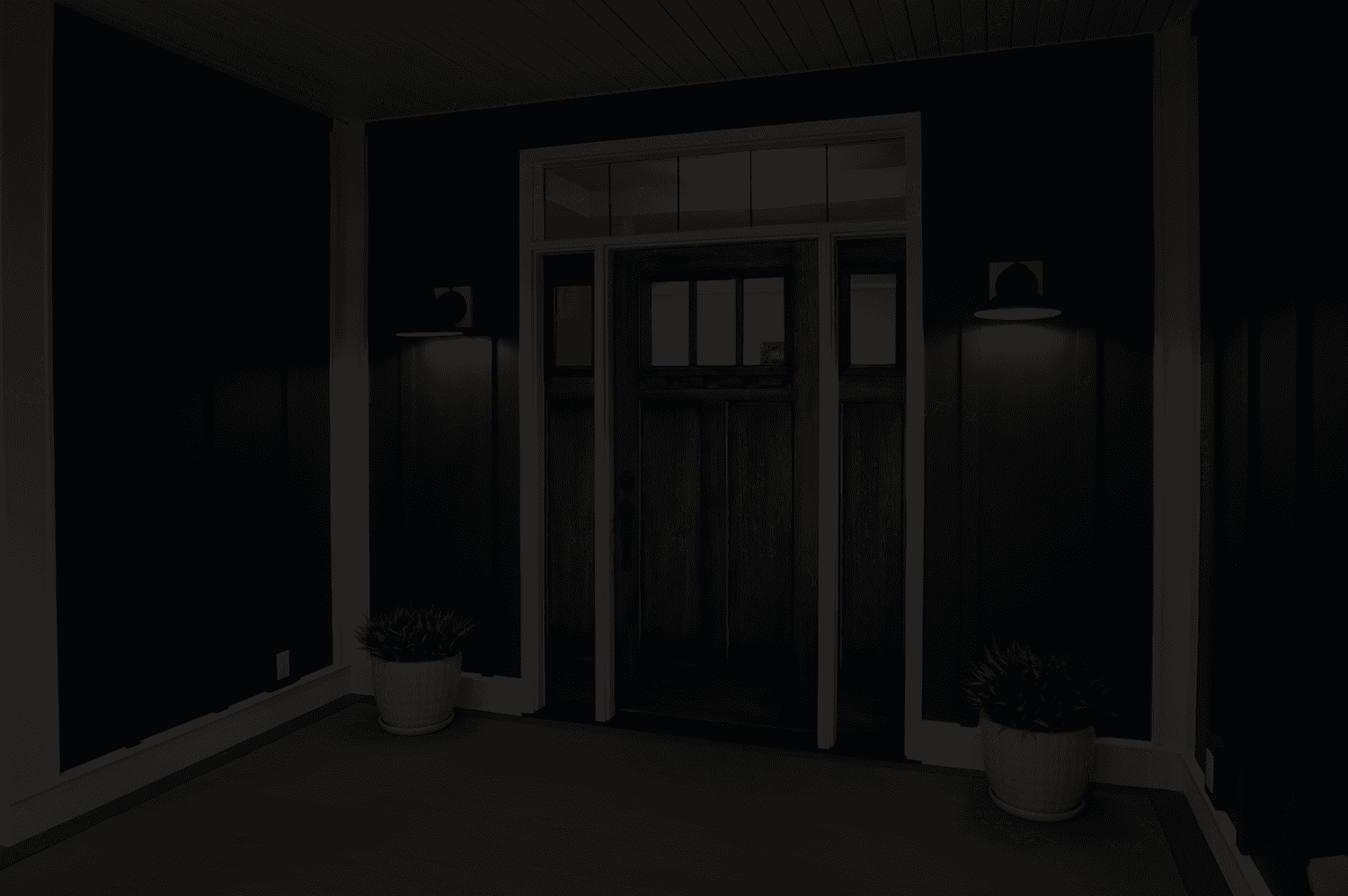 A dark, elegant front porch with navy blue walls, illuminated by two black wall sconces flanking a wooden door. Two potted plants add a welcoming touch.