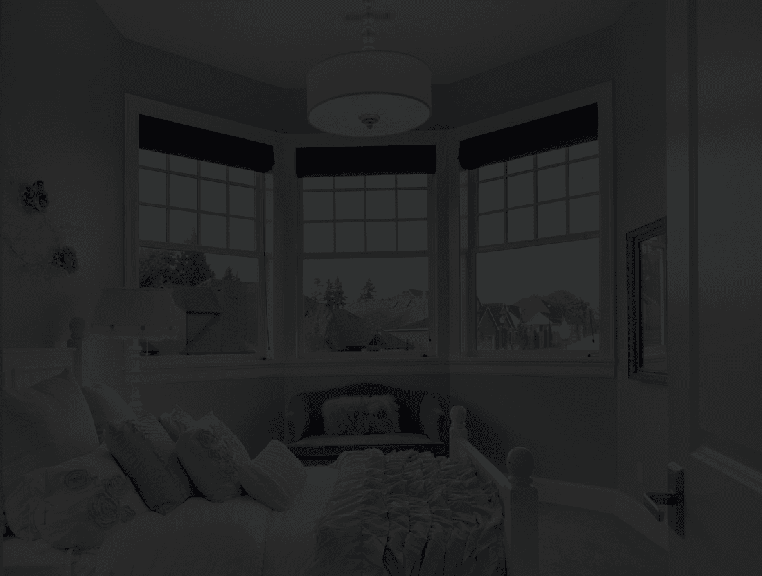 A cozy bedroom with a soft blue theme features a bed with white and blue pillows, a blue loveseat with a pink pillow, and a large bay window with dark blinds.