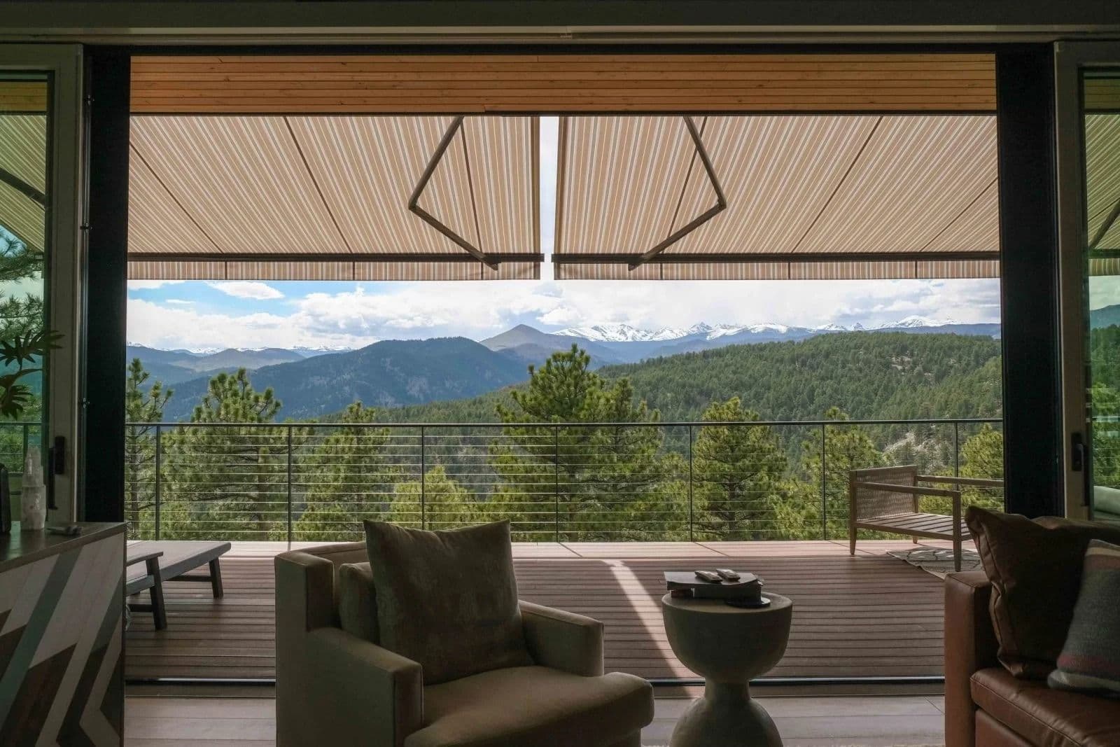 Modern living room with large windows opens to a scenic mountain view. Cozy chairs and a table inside, with greenery and snowy peaks outside, creating a serene atmosphere.