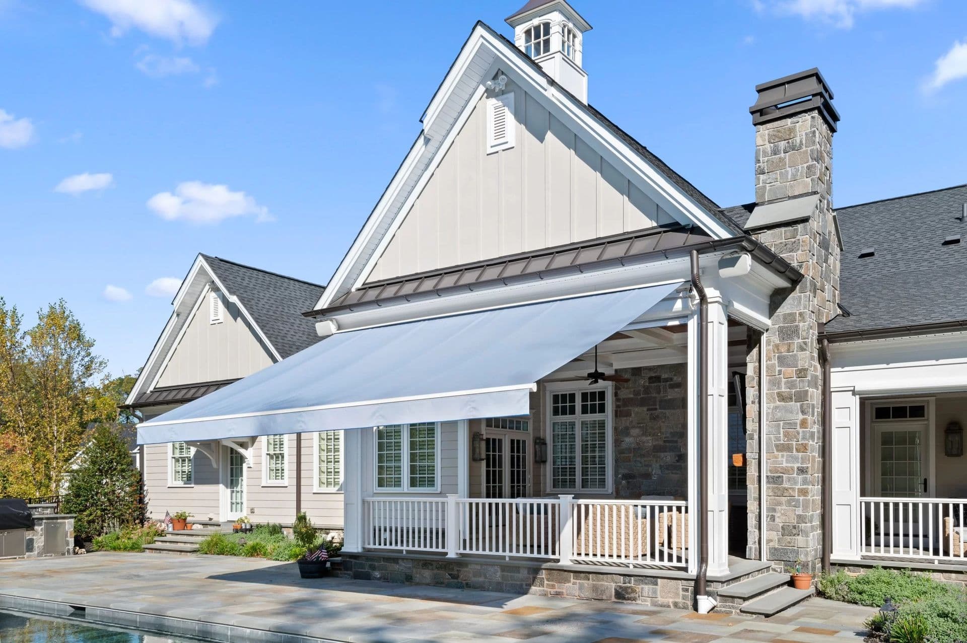 A modern house with gray siding, a stone chimney, and a large white awning over a porch. The atmosphere is sunny and serene, with a clear blue sky.