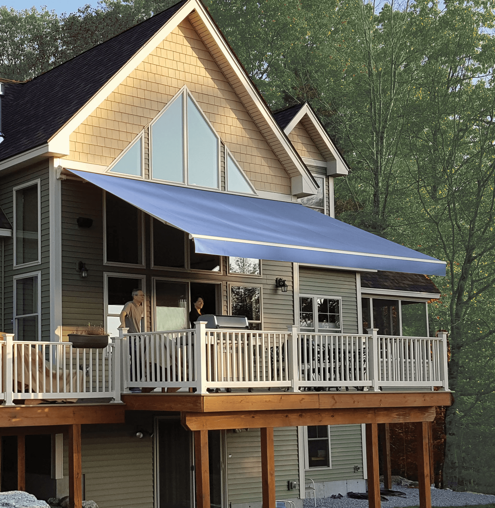 A modern house with a tall, triangular roof and large glass windows. A blue awning covers the balcony where two people stand, surrounded by trees.
