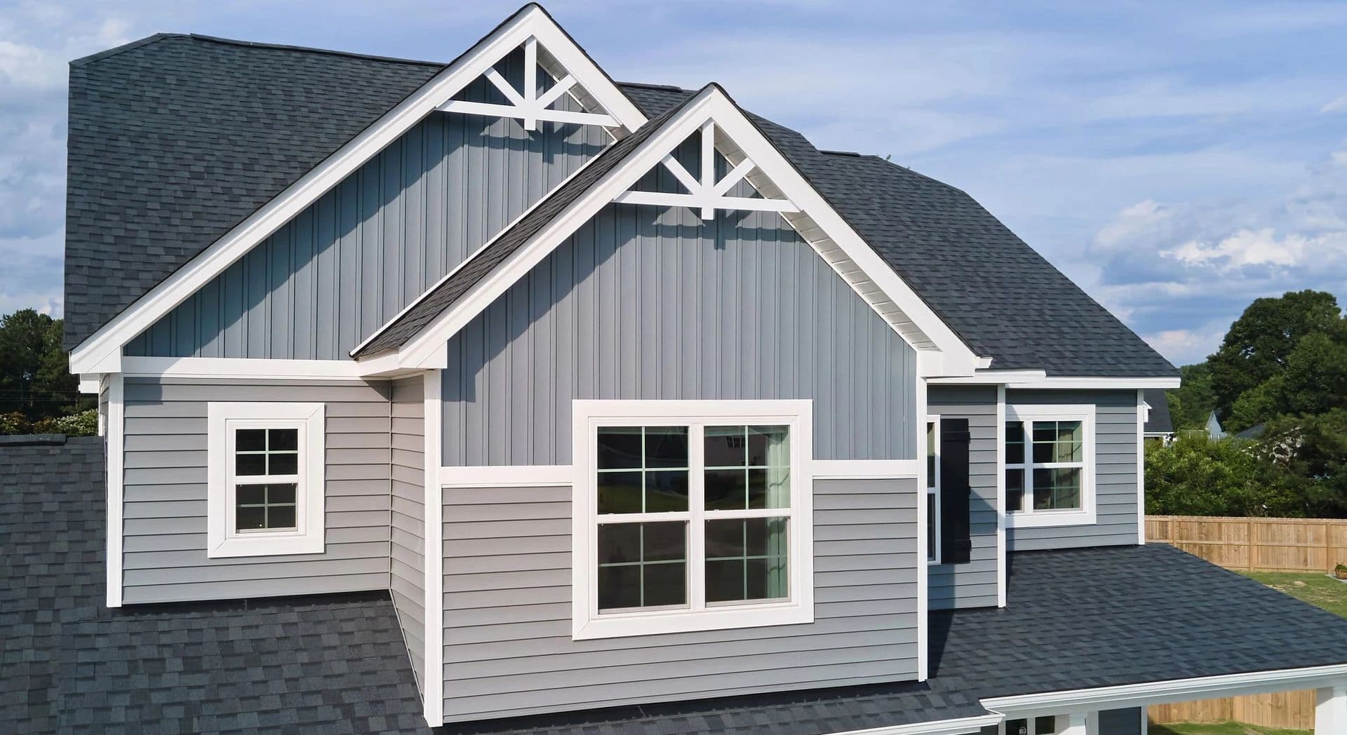 Gabled roof house with gray siding and white trim, featuring large and small windows. The sky is blue, conveying a calm suburban setting.