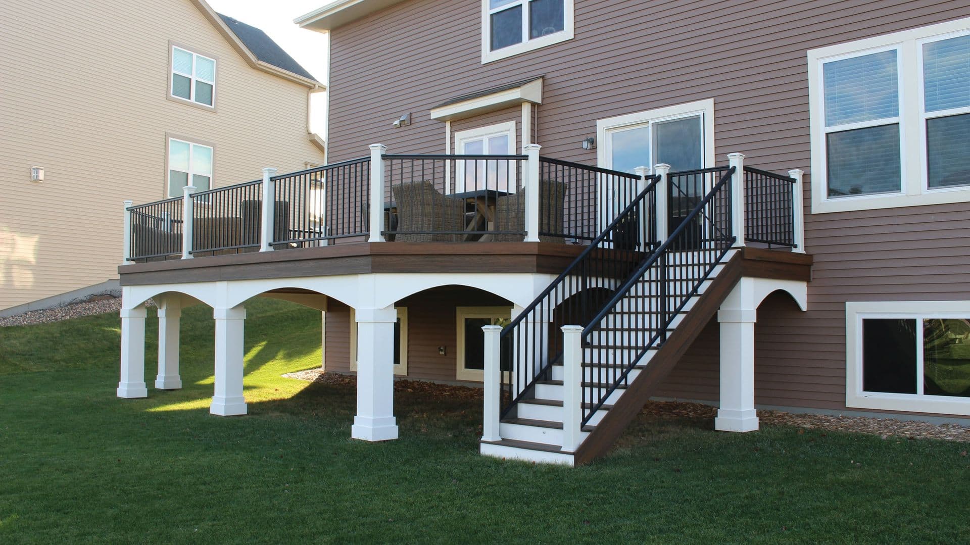 A large brown house with a raised wooden deck, white pillars, and black railings. The deck has stairs leading down to a green lawn on a sunny day.