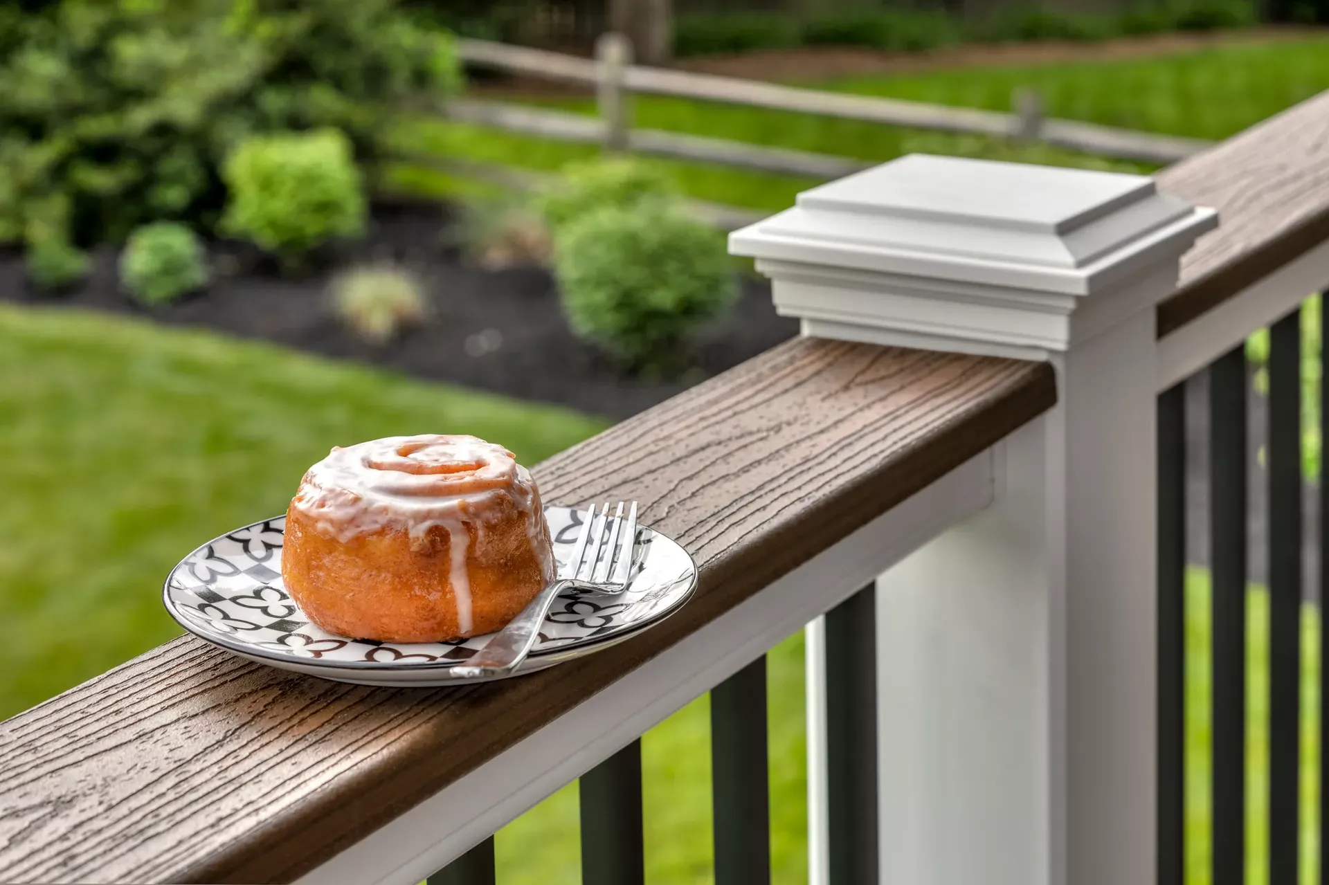 A glazed cinnamon roll on a decorative plate with a fork, resting on a wooden deck railing. Lush garden greenery and a fence are in the background.