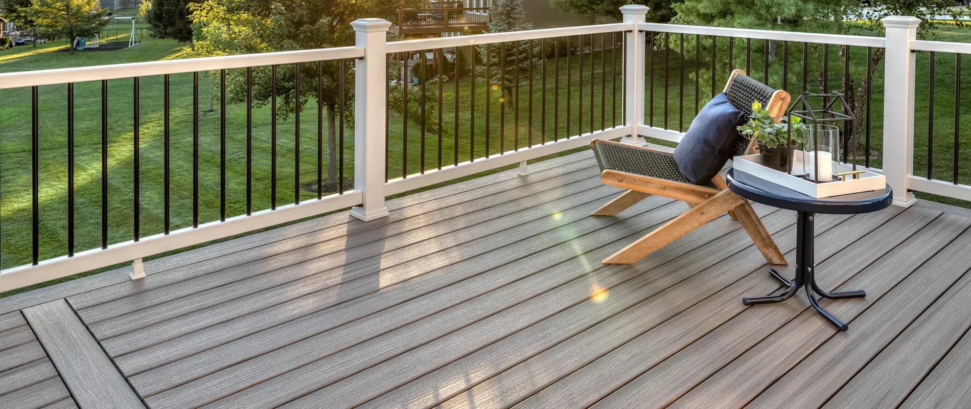A spacious wooden deck overlooks a green lawn at sunset. It features a simple chair with a cushion and a small round table holding a lantern, creating a serene ambiance.