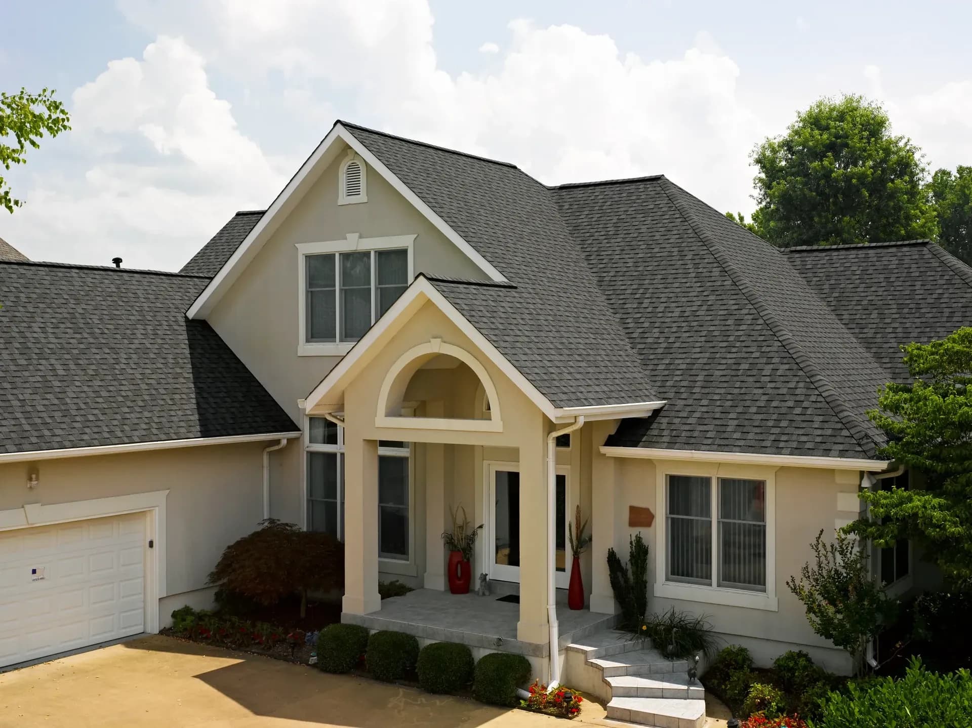 Spacious modern house with dark shingle roof, arched entrance, and two-story windows. Manicured shrubs frame the steps. Bright, sunny day.