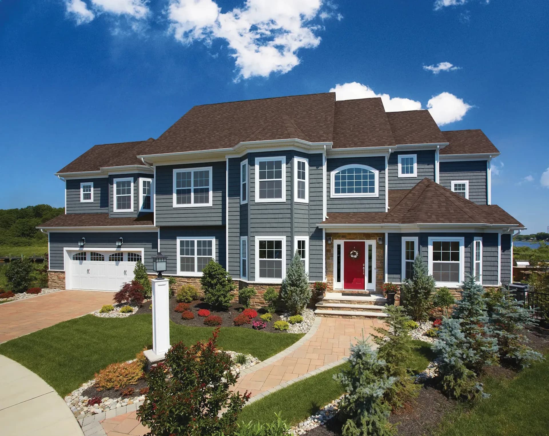 A stately two-story blue house with white trim and a red front door. The landscaped yard features bushes and trees, set under a bright blue sky.