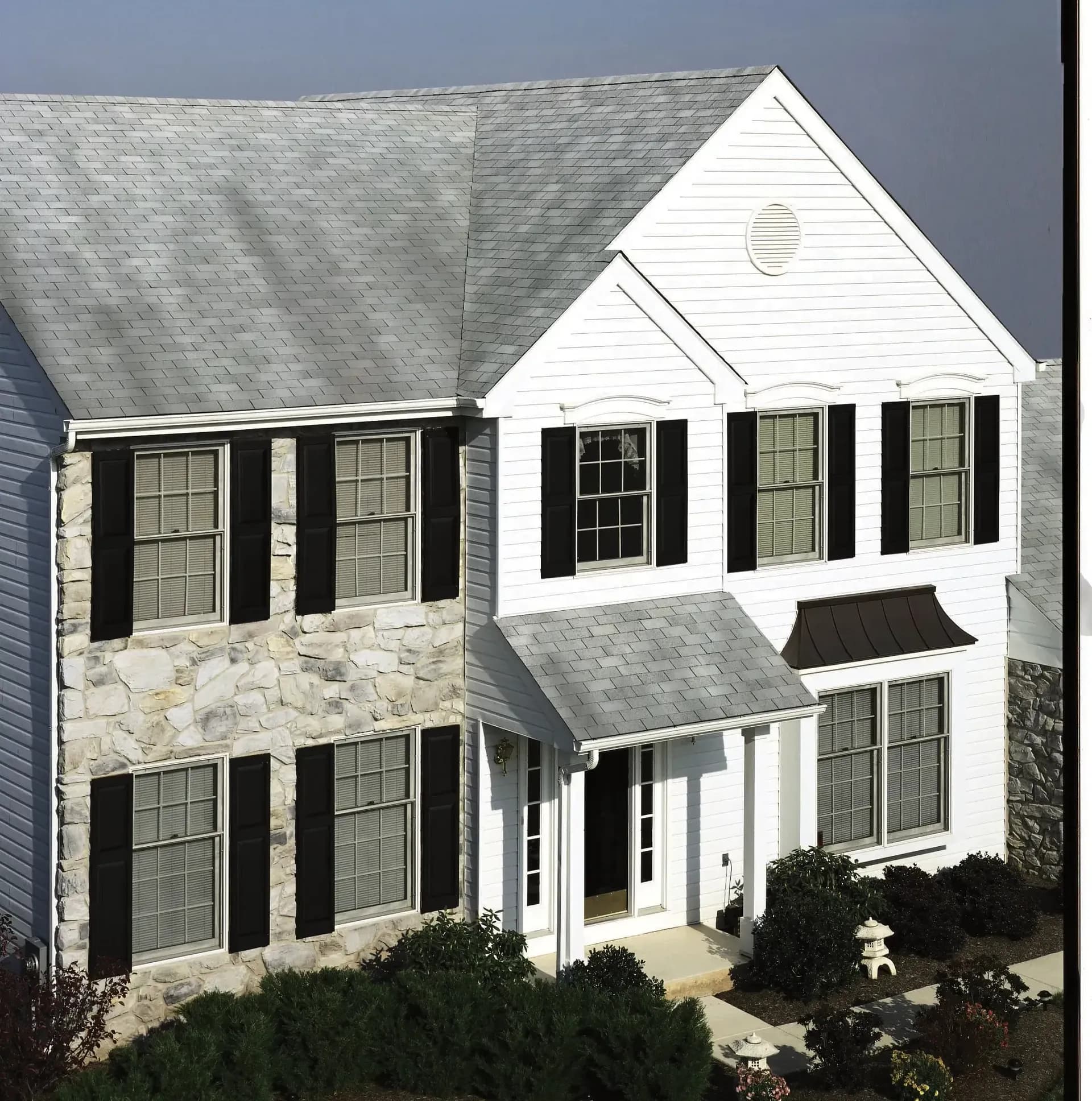 A two-story house with white siding, stone accents, and dark shutters. It has a gray gable roof and a landscaped garden, conveying a neat, suburban feel.