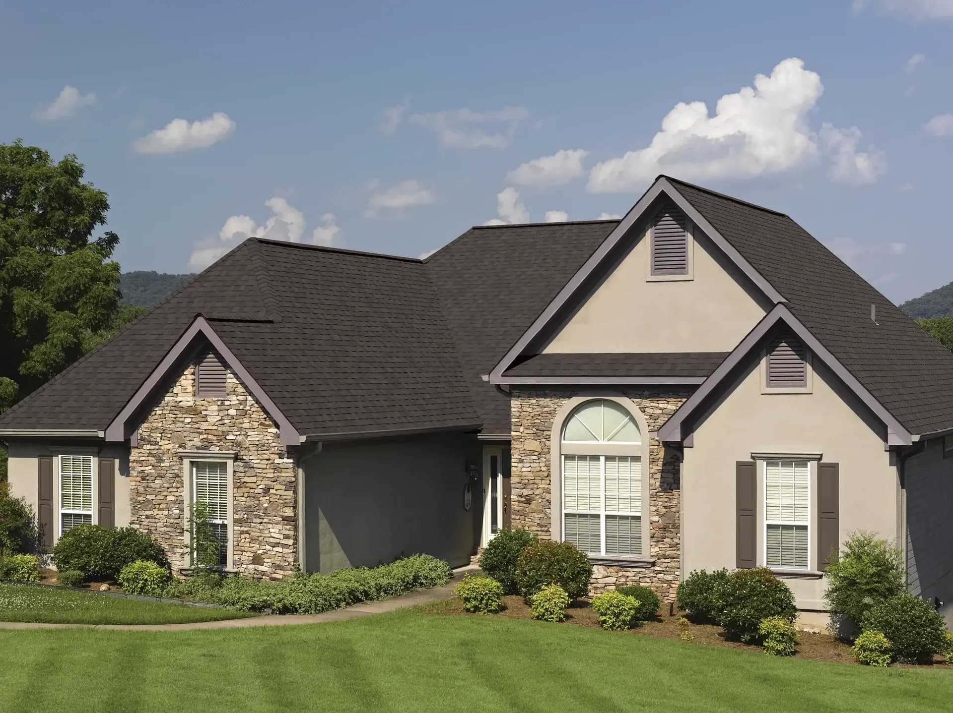 Single-story house with gray siding and stone accents, dark roof, and arched window. Surrounded by green lawn, bushes, and trees under a clear blue sky.