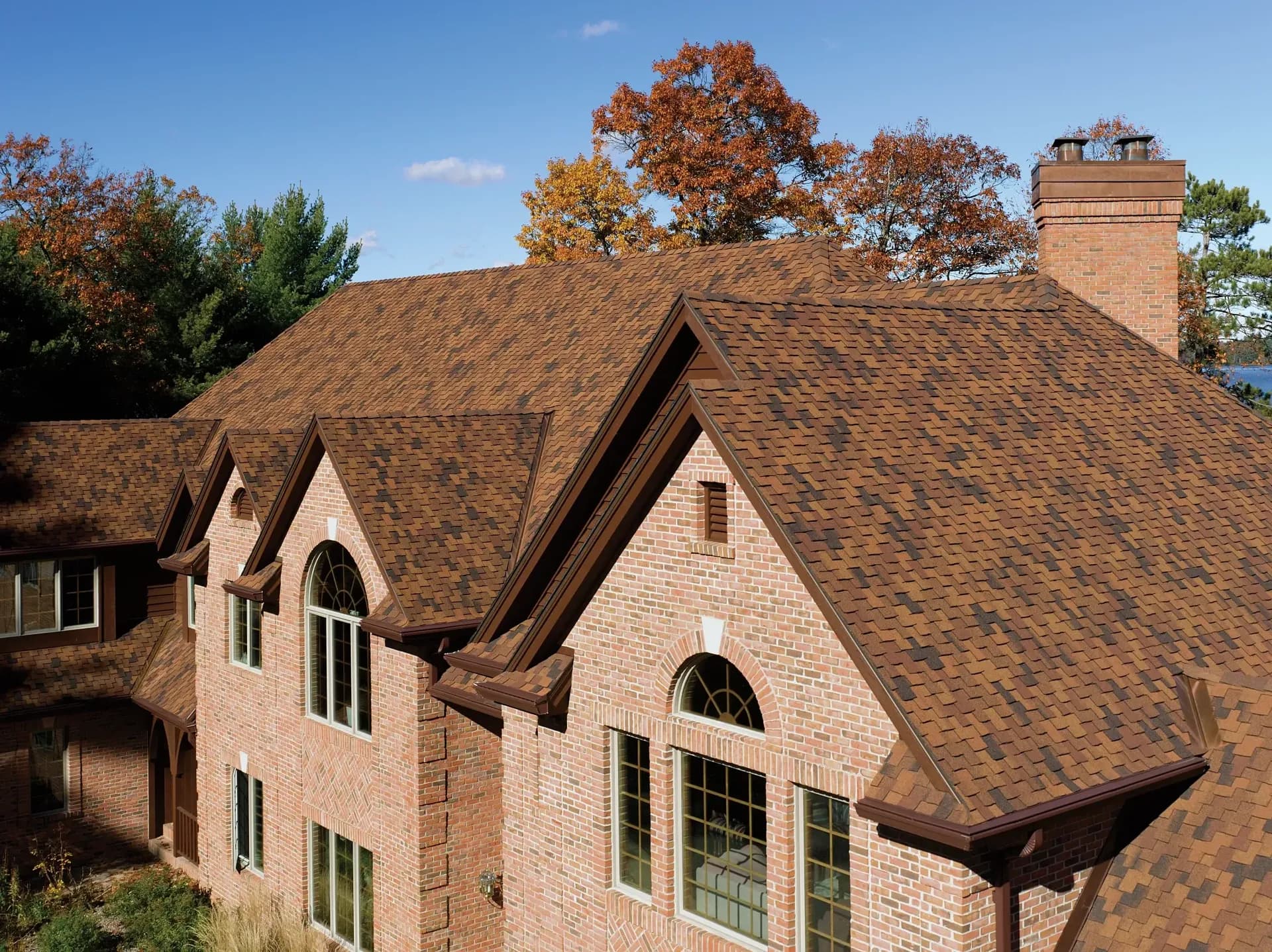 Red brick house with brown shingle roof, surrounded by autumn trees. Blue sky in the background creates a warm, inviting atmosphere.