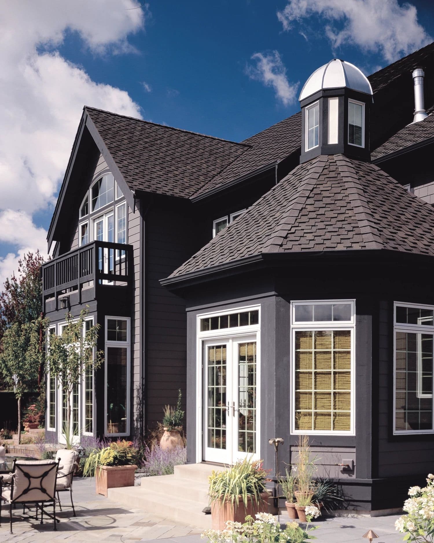 Modern gray house with a steeply pitched roof, expansive windows, and a small balcony. The sky is clear, creating a peaceful, sunny ambiance.