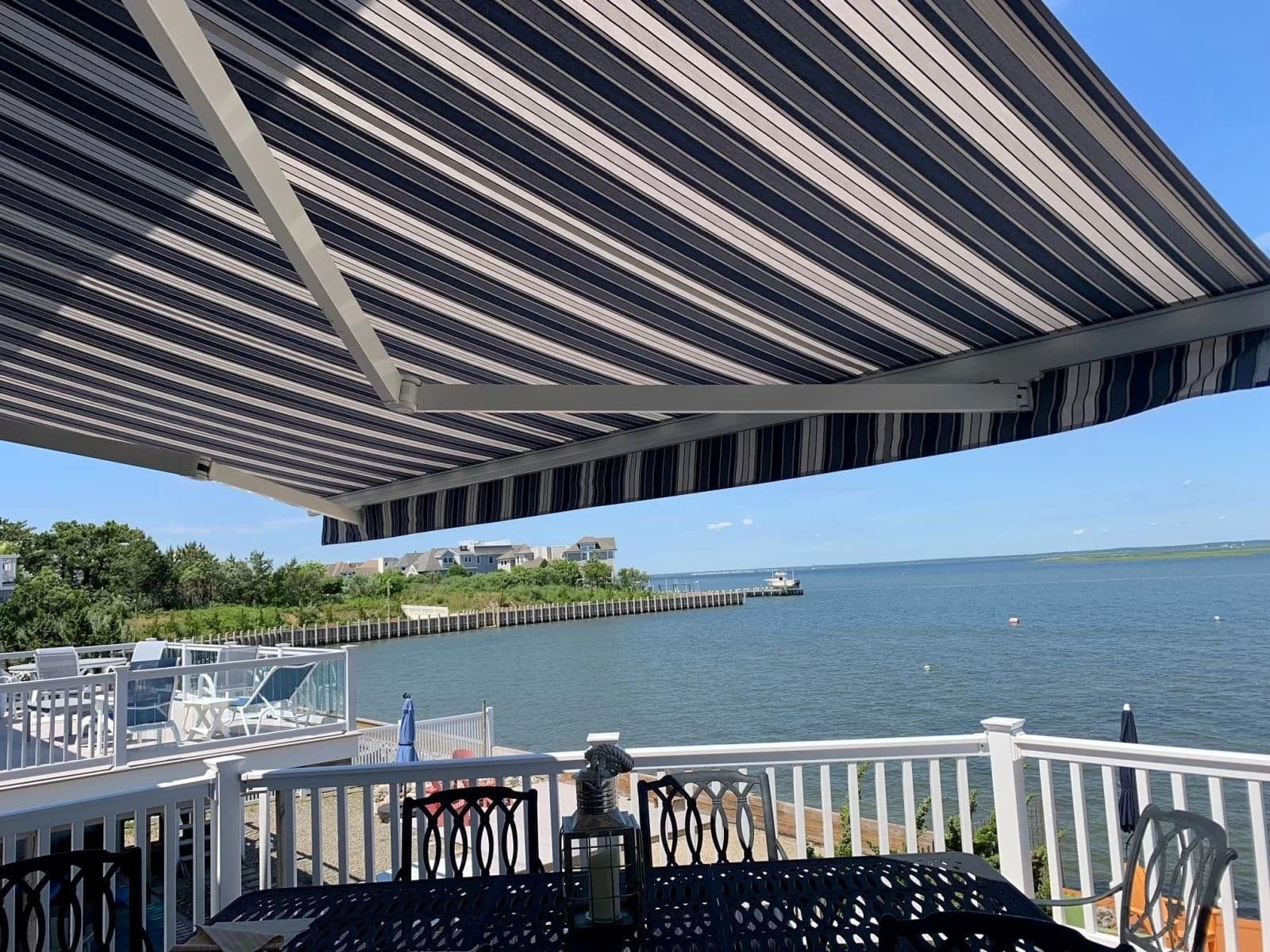 Striped awning over a patio with black metal furniture, overlooking a serene waterfront view. Blue sky and trees create a relaxing, coastal atmosphere.