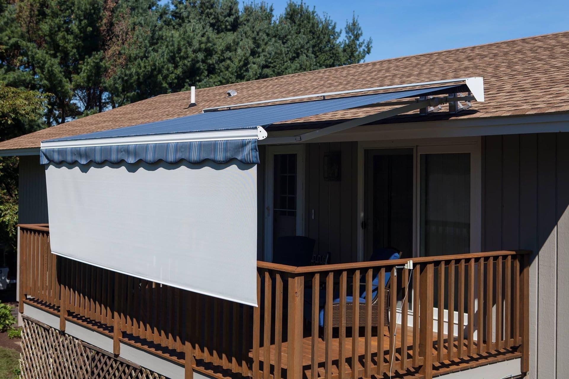 Rooftop balcony with wooden railing, featuring a retractable awning and white fabric sunshade; trees in the background under a clear blue sky.