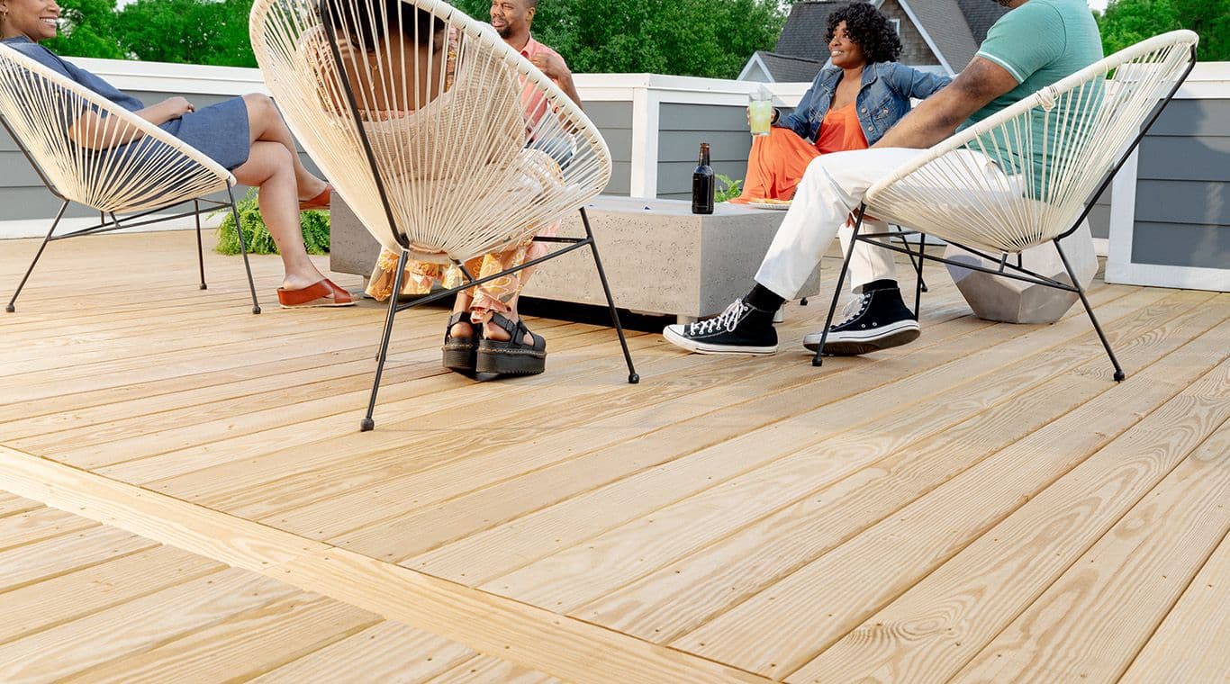 Group of people sitting on modern chairs around a low table on a wooden deck. They are relaxed, enjoying drinks, with trees and houses visible in the background.