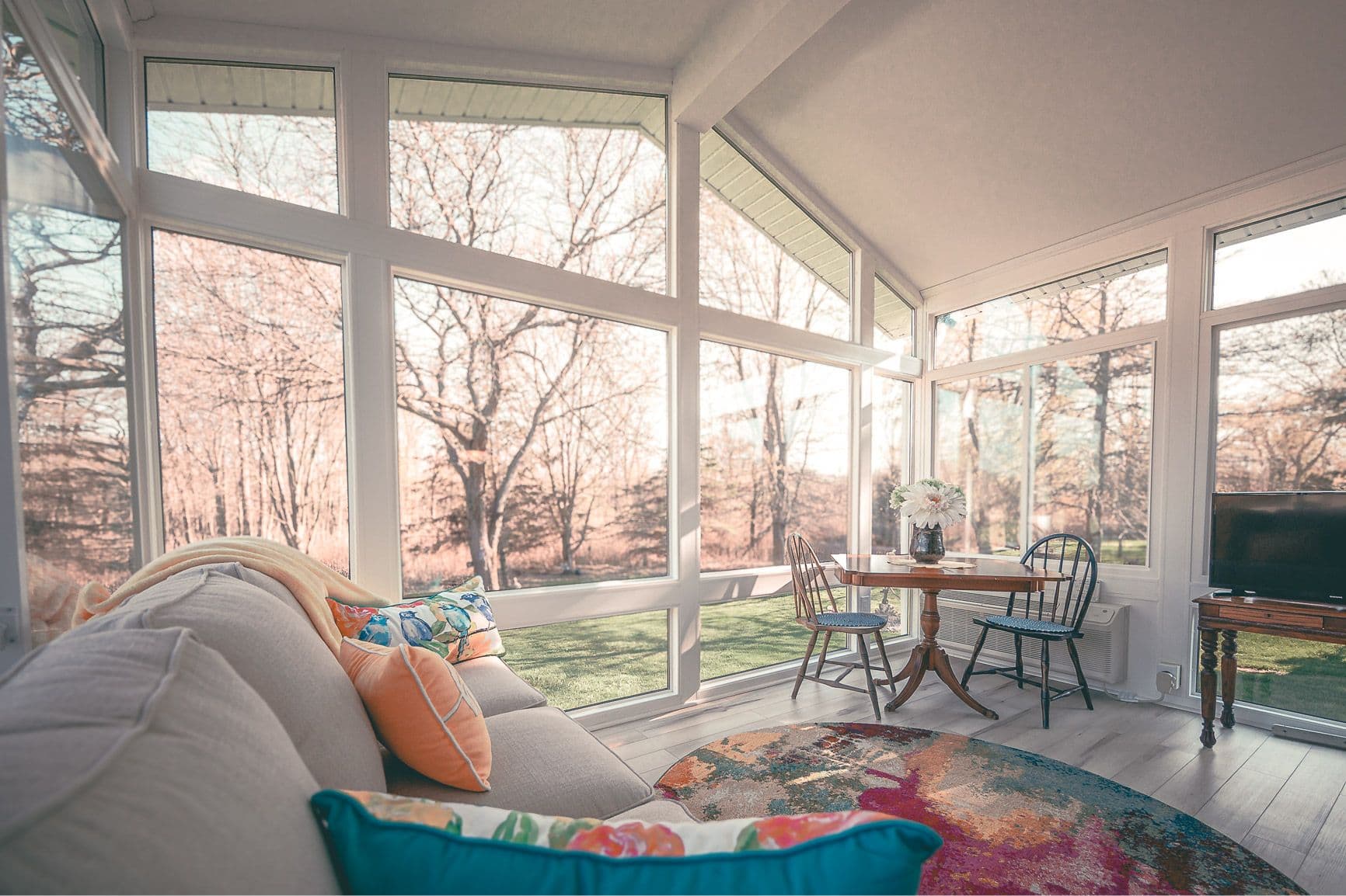 Bright sunroom with large windows showcasing a wooded view. A cozy sofa with colorful pillows and a round table set for two creates a serene ambiance.