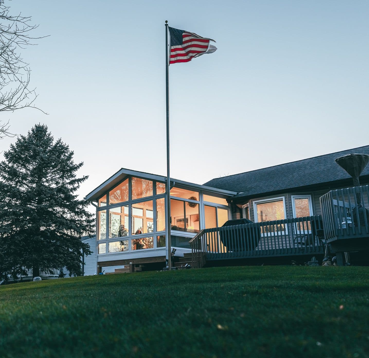 A U.S. flag flutters on a pole beside a modern house with large windows glowing warmly at dusk. A tree stands nearby, and the lawn is lush and green.