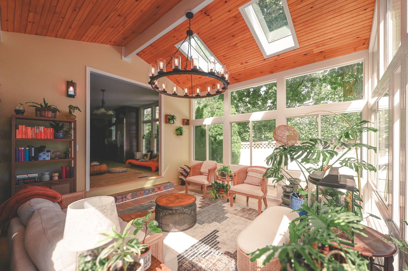 A sunlit room with large windows and a wooden ceiling features cozy seating, lush plants, and a chandelier. The space conveys warmth and relaxation.