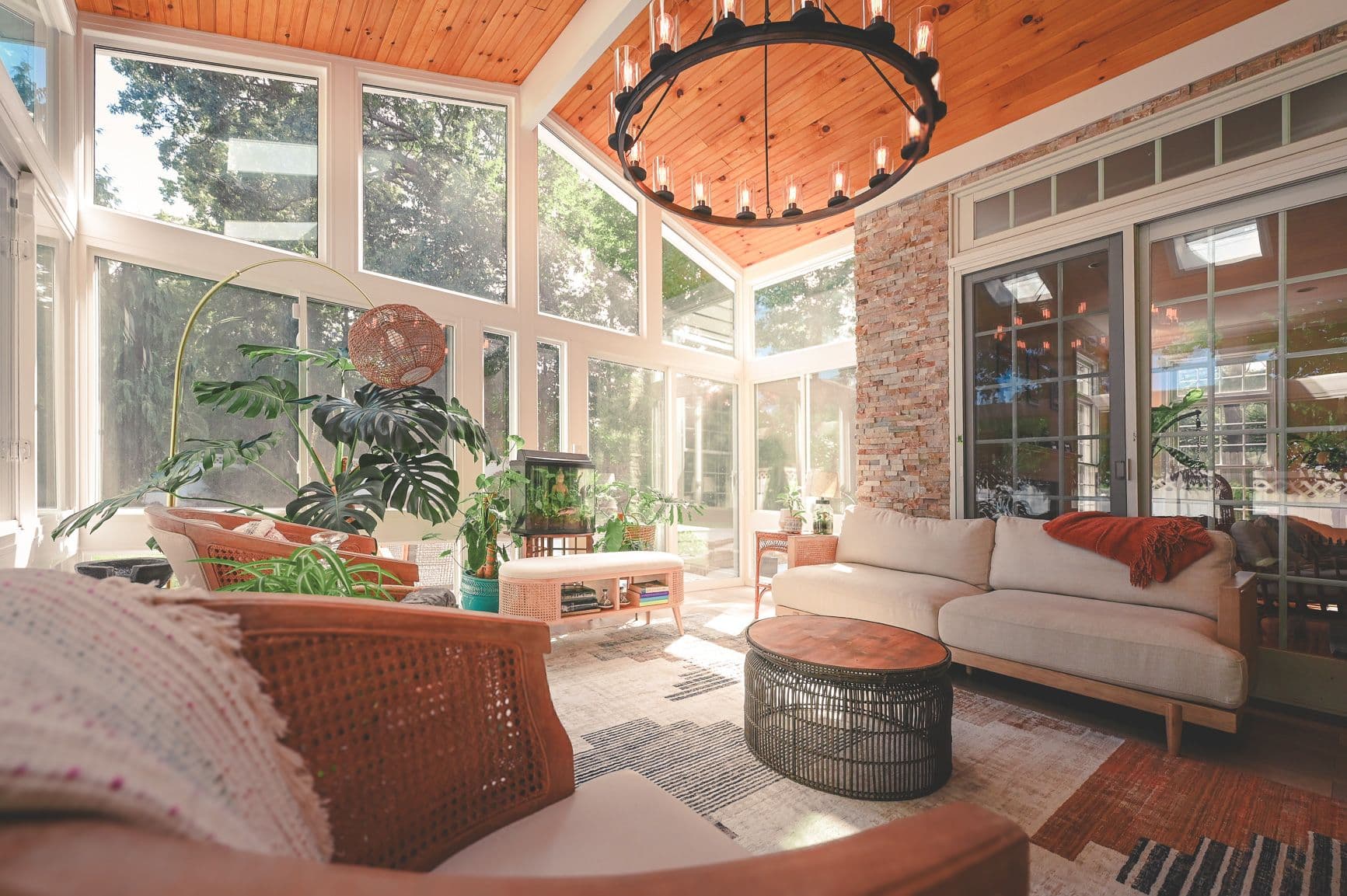 A cozy sunroom with abundant natural light, featuring a plush sofa, wooden coffee table, wicker chairs, lush green plants, and a large chandelier.