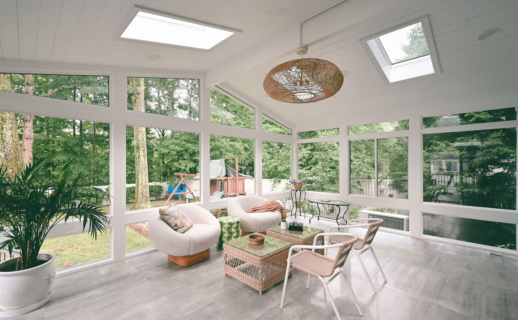 Bright sunroom with large windows showcasing a lush green garden. Features two cozy armchairs, a wicker table, a potted plant, and rattan ceiling light, conveying a serene, airy atmosphere.