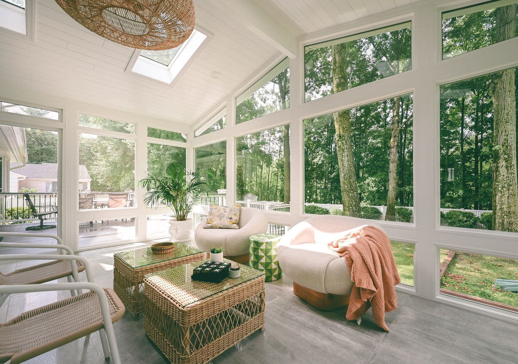 Bright sunroom with large windows overlooking a forest. Includes cozy chairs, wicker coffee table, and a plant, creating a tranquil, inviting atmosphere.