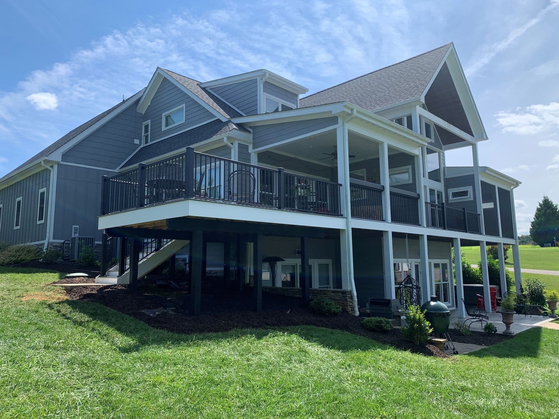 Modern house with gray siding and a covered deck, elevated on stilts. Sunny day, with green grass and blue sky in the background, conveying a serene atmosphere.